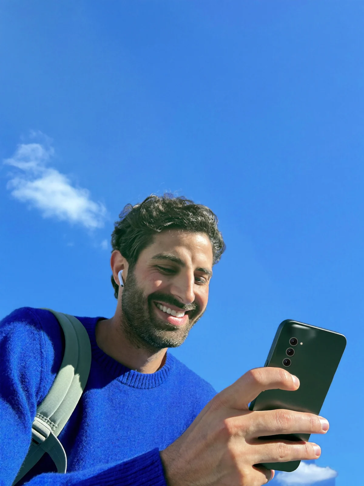 Un homme en pull bleu sourit en lisant son téléphone, sur fond de ciel